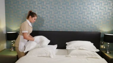 Profesyonel temizlik servisi. Yatağı toplamaktan, dövmekten ve yastıkları ziyaretçiler için hazırlamaktan gülümseyen oda hizmetçisi. Yastıkları kabartan kadın. 4k video