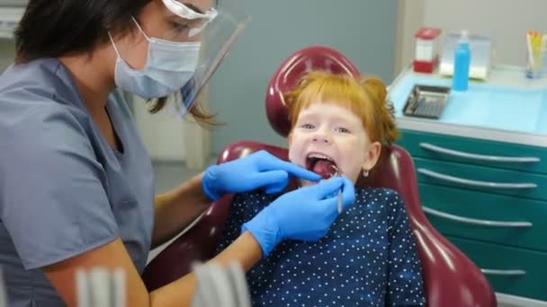 Odontología pediátrica. Primer plano de la dentista que trata a la niña pelirroja en la clínica dental moderna. Un niño encantador en el consultorio del dentista. 4 k vídeo — Vídeo de stock