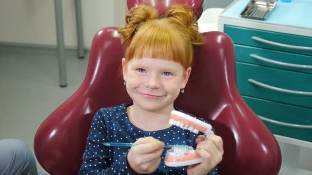 Pediatric dentistry concept. Charming little girl in dental chair. Lovely red-haired girl having fun at dental office, playing with teeth model cleaning teeth with brush. 4 k video — Stock Video