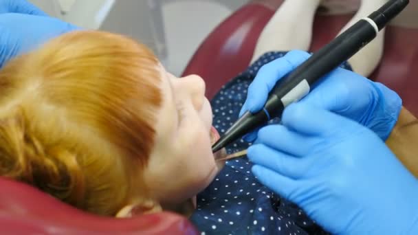 Детская стоматология. Крупным планом женщина-стоматолог лечит маленькую рыжую девочку в современной стоматологической клинике. Прекрасный ребенок в кабинете дантиста. 4 k видео — стоковое видео