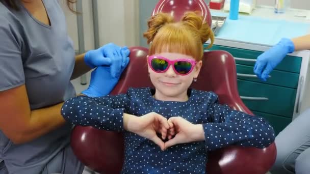 Un niño sonriente y seguro en el consultorio del dentista. niña feliz con gafas de sol sentado en la silla dental que muestra los pulgares hacia arriba después de la revisión dental. Odontología pediátrica. 4 k vídeo — Vídeo de stock