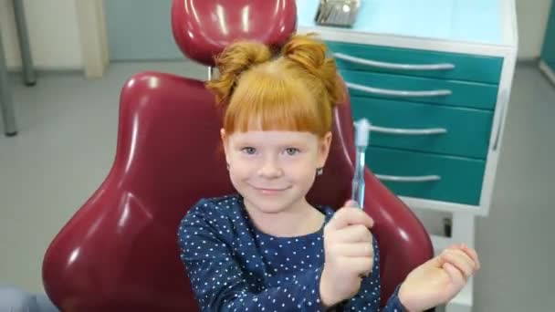 Gyermekfogászat koncepció. Elbűvölő kislány fogorvosi székben. Kedves vörös hajú lány, aki jól érzi magát a fogorvosi rendelőben, fogkefével játszik. 4 k videó — Stock videók