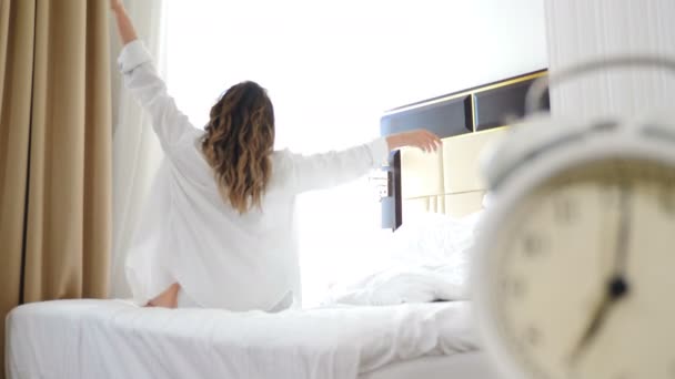 Portrait of pretty young woman in white shirt sitting on bed in front of large window lit with sunshine. Shot through white alarm-clock. Morning concept. 4 k video — Stock Video