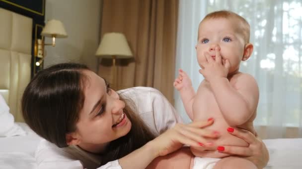 Gelukkig zwangerschapsconcept. liefdevolle jonge moeder knuffels verpleging 8 maanden dochter op sneeuw-witte deken in gezellige slaapkamer. tederheid en geluk van het gezin. kinderen, kind, zuigeling, kind, baby. 4 k video — Stockvideo