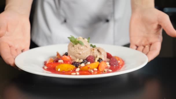 Concetto di ristorazione. chef che presenta piatto di cibo con delizioso dessert alla frutta in stile flambe. Primo piano. Ristorante cucina e servizio. Mani dello chef che tengono il piatto bianco e si mettono sul tavolo nero — Video Stock