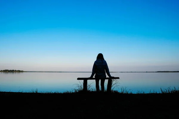 Silhouette of a meditating woman 59 years old at dawn on Svityaz Lake in Ukraine. The woman is sitting on a home-made wooden bench.