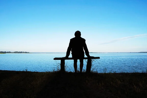 Silhouette of a meditating man 58 years old at dawn on Svityaz Lake in Ukraine. The man is sitting on a home-made wooden bench.