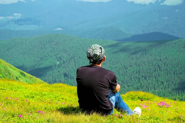 Happy man 58 years old sits among the colors of rhododendron in the alpine meadows in the Carpathian Mountains. View from the back. Ukrainian tourism.