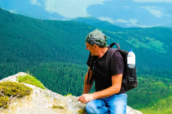A tourist with a backpack sits on a stone high in the mountains. In the backpack pocket is a water bottle mock up. Ukrainian tourism in the Carpathian Mountains.