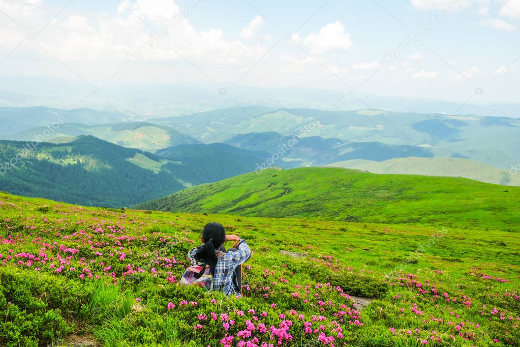Happy woman 59 years old sits among the flowers of Rhododendron in the Alpine meadows in the Carpathians mountains. Ukrainian tourism. View from the back.