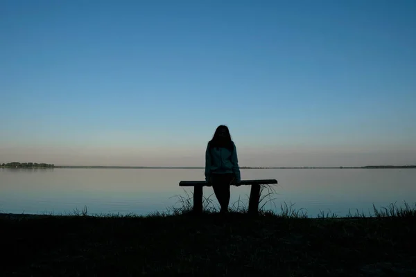 Silhouette of a meditating woman 59 years old at dawn on Svityaz Lake in Ukraine. The woman is sitting on a home-made wooden bench.