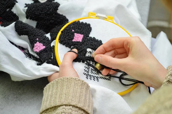 The woman embroiders by hand embroidery on white fabric with black and pink wool threads in hoop, left view, macro