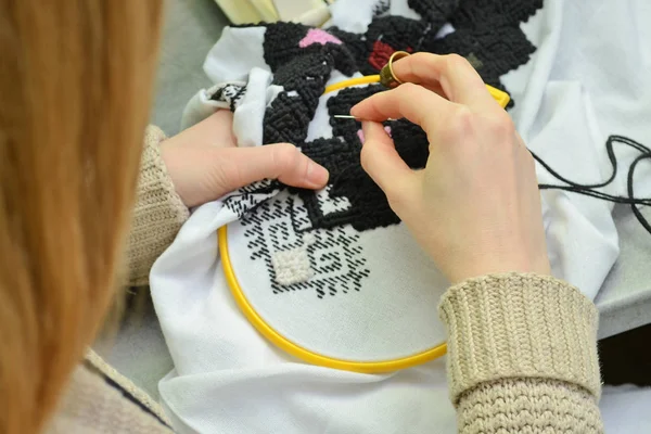 The woman embroiders by hand Ukrainian embroidery on white fabric with black and pink wool threads in the hoop, closeup