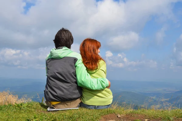 Happy white man and woman tourists in green clothes sitting hugging on edge of top of mountain and look into distance