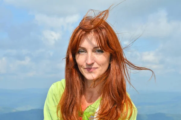 Happy smiling redhead woman tourist in the sunlight in the mountains against the sky, the wind ruffles her hair