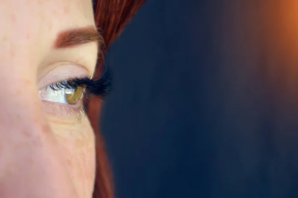 Oog van het meisje met rood haar en groene ogen met wimper Extensions op een donkere achtergrond ziet er links uit met gloed en copyspace — Stockfoto
