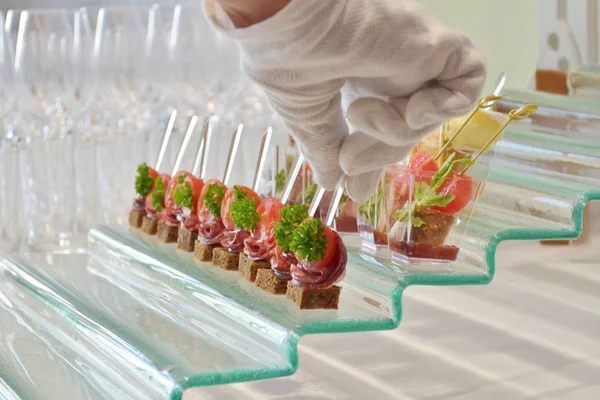 Verschiedene Arten von Canapes mit transparenten Spießen auf einem gläsernen Stufenständer auf dem Buffettisch, eine behandschuhte Hand legt sie aus — Stockfoto