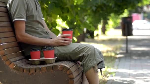 Bir adam ağaç altında sokakta ahşap tezgah üzerinde oturur ve kırmızı fincan kahve içmek — Stok video