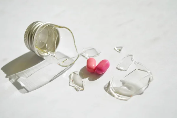 Ένα σπασμένο μικρό γυάλινο μπουκάλι με μεταλλικό καπάκι, δύο ροζ δισκία σε σχήμα καρδιάς έπεσαν από αυτό, σε βρώμικο λευκό φόντο — Φωτογραφία Αρχείου