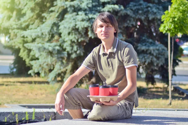 Young white man sits on a concrete parapet in a park and holds four red paper glasses with coffee