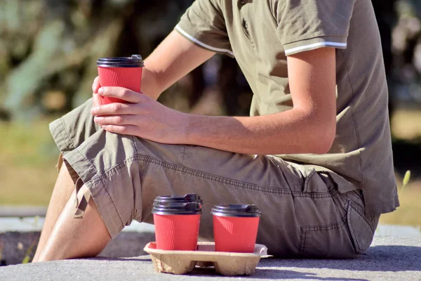 A young white man with no face sits on parapet in park, near him are red paper glasses with coffee and one in his hand