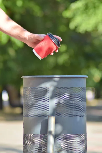 Το χέρι ενός άντρα ετοιμάζεται να πετάξει ένα κόκκινο χάρτινο κύπελλο για καφέ σε ένα κάδο σκουπιδιών, με προσανατολισμό — Φωτογραφία Αρχείου