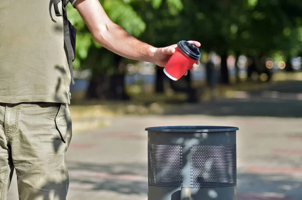Rahat bir tarzda Adam yeşil ağaçlar ve şehir bir arka plan üzerinde, bir çöp konteyner üzerinde kahve için kırmızı kağıt fincan tutar