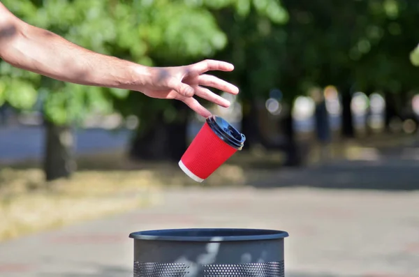 Erkek el bir çöp konteynerkahve için kırmızı bir kağıt fincan attı, fincan havada sinekler, yeşil ağaçların arka plan