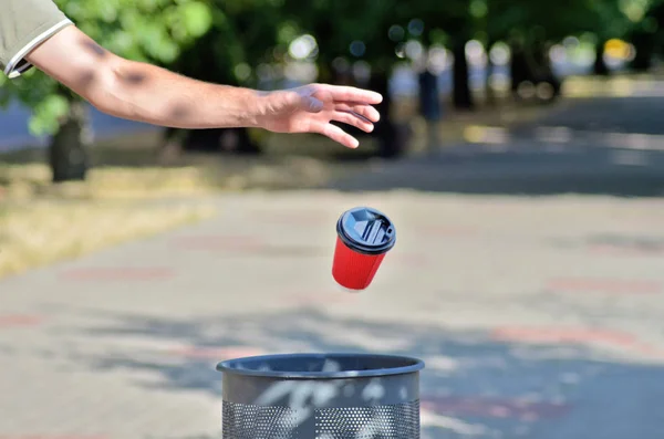 Erkek el bir çöp kahve için kırmızı bir kağıt fincan attı, fincan havada sinekler, yeşil ağaçlar ve sokak arka plan karşı