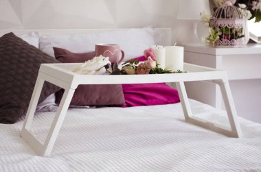 Üzerinde bardak, çiçek ve mumlar olan beyaz bir yatakta beyaz bir kahvaltı masası. Yakında pembe ve kahverengi yastıklar var.