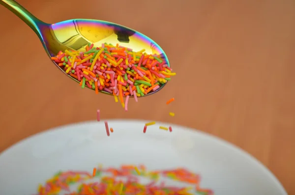 Long baking topping, sprinkled from a rainbow spoon in a white saucer, on a wooden table — Stock Photo, Image