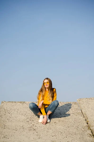 stock image Beautiful blonde girl in yellow sweater and jeans, sneakers sits on concrete surface against blue sky in yellow glasses