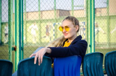 Sarı kazaklı, beyaz spor ayakkabılı ve mavi ceketli güzel sarışın kız spor sahasındaki seyirciler için koltuklarda oturuyor.