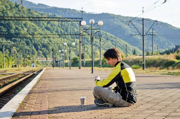 Joven con ropa de turista se sienta en la plataforma de la estación, mira el reloj mientras espera el tren, con una taza de café — Foto de Stock