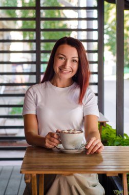 Genç Avrupalı kız, yaz mevsiminde elinde fincanla tahta masada, çizgili arka planı ve çiçekleri olan bir kafede oturuyor.