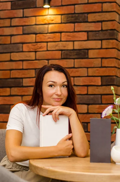 Młoda szczęśliwa biała kobieta z brązowymi włosami siedząca w kawiarni przy ceglanej ścianie z książką przy stole — Zdjęcie stockowe