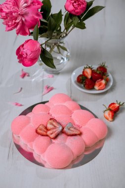 Çilekli kremalı pasta masanın üzerinde duruyor. Bir buket pembe şakayık ve bir dilim çilek..