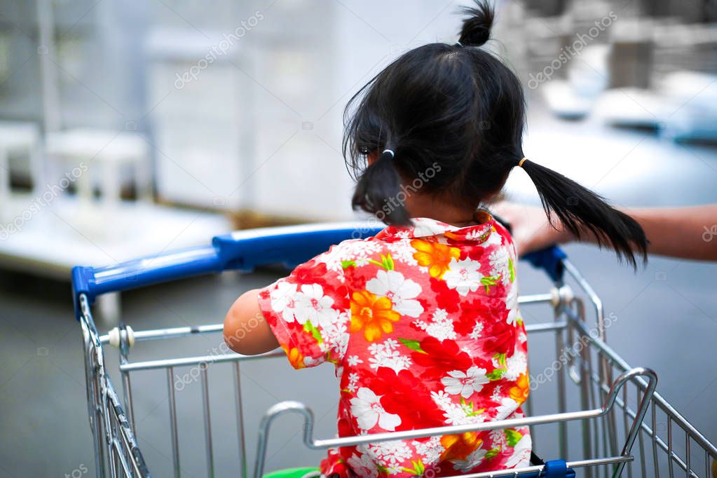 Asian little girl in orange flower shirt sits in supermarket cart and spins around her head in front between shopping furniture in the warehouse shopping center.