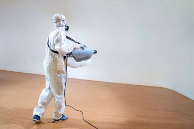 Önleme giysisindeki profesyonel teknik adam, elektrikli sprey makinesiyle tahta zemine sterilizasyon solüsyonu sıkıyor ve beyaz arka planda stüdyo ışığı var..