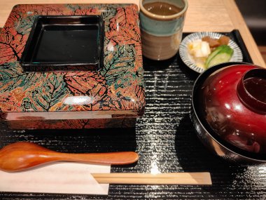 Klasik bir Japon setinde çorba, turşu ve sıcak çay eşliğinde ızgara yılan balığıyla servis edilen laktüel bir kutu yemek. Sunumda denge ve lezzete dikkat ederek geleneksel akşam yemeklerine vurgu yapılıyor.