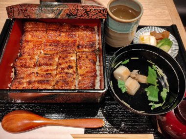 Klasik bir Japon setinde çorba, turşu ve sıcak çay eşliğinde ızgara yılan balığıyla servis edilen laktüel bir kutu yemek. Sunumda denge ve lezzete dikkat ederek geleneksel akşam yemeklerine vurgu yapılıyor.