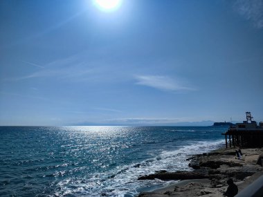 Dalgalar parlak mavi bir gökyüzü altında kayalık kıyıya hafifçe çarpar ve güneş ışığı denizi yansıtır. Kıyı manzarası okyanusun ve ufkun doğal güzelliğini vurgular..