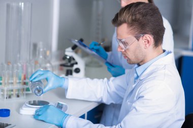 Genç ciddi sorumlu araştırmacı oturan ve madde üzerinde ölçekler ağırlığında laboratuarında çalışan bir gözlük ve lastik eldiven.