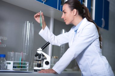 Uzun zaman bekledi sonucu. Madde ile şişe tutmak ve mikroskop kullanarak laboratuar bir üniforma ayakta genç nitelikli sorumlu uzman.