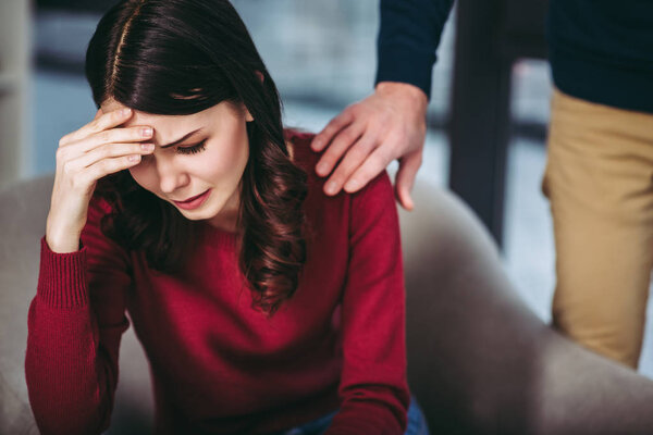 portrait of depressed woman sitting in the armchair and man holding hand on her shoulder