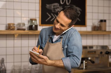 kendine güvenen kafe sahibi Not almak gülümseyen ve Smartphone'da konuşuyor