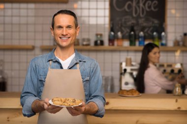 kruvasan plaka üzerinde tutarak ve arka planda garson ile kameraya bakarak garson gülümseyerek