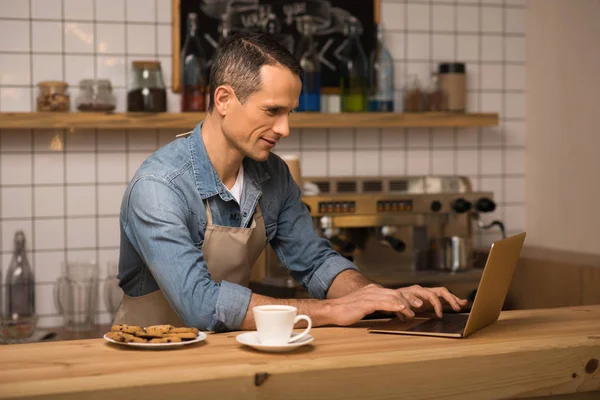 Genç barista dizüstü kullanarak önlük yan görünüm