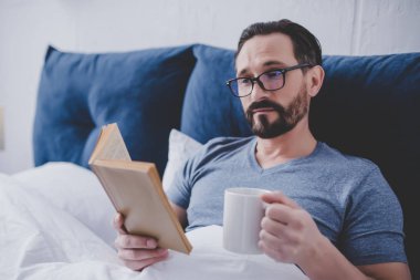 Adam kitap ve holding fincan çay yatakta gönüllülük gözlük