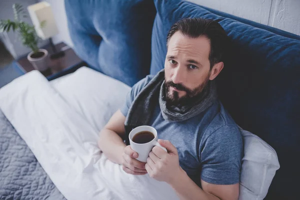 ill man wearing woolen scarf and drinking tea sitting in the bed at home, looking at camera
